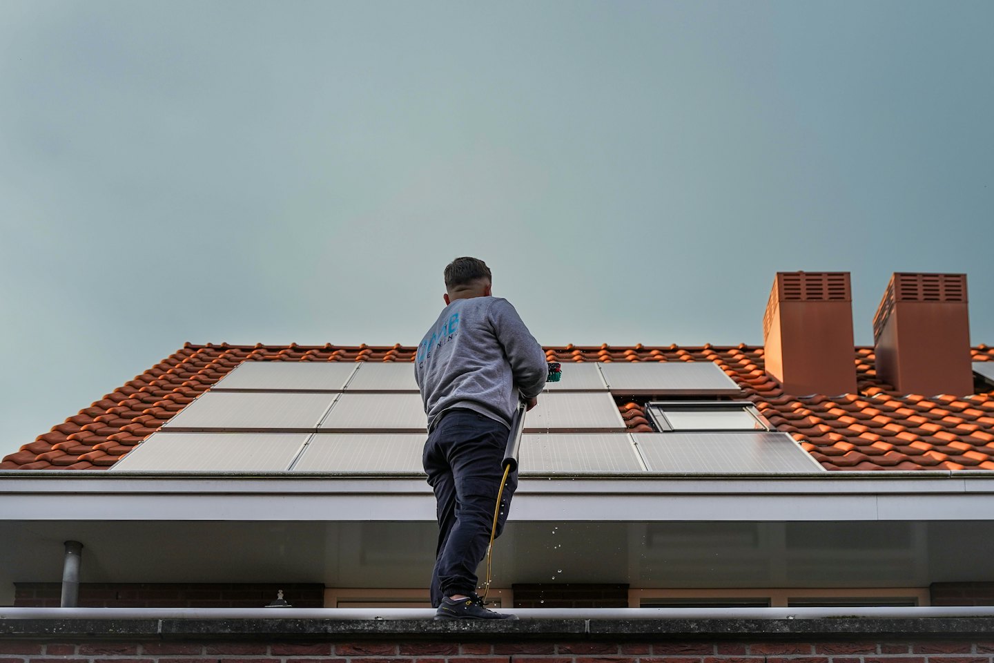 Pracownik czyści panele słoneczne na dachu z pomarańczowej dachówki długą szczotką.